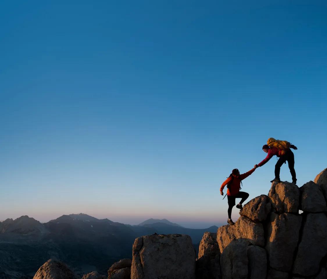 Photo: atop a mountain at dusk; two hikers, one helping the other reach the top