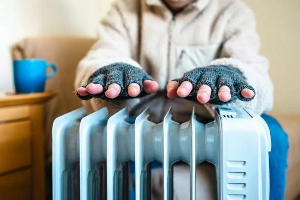Image of someone heating themselves on a small radiator with gloves and a coat on. Everything you need to know about the cold-health alert