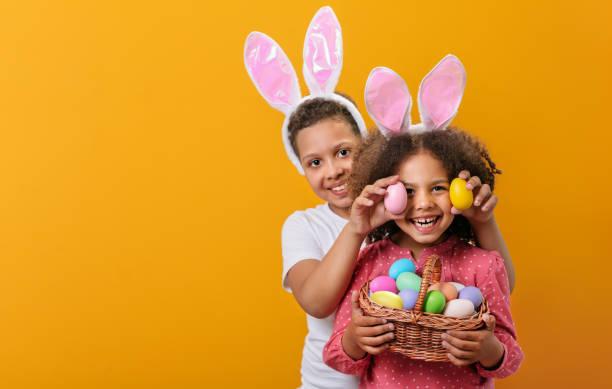 two children wearing bunny ears holding a basket of multi-coloured Easter eggs