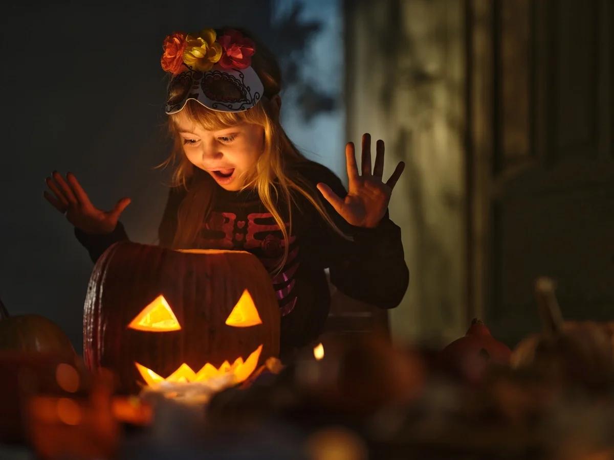 A little girl in fancy dress looks in open-mouthed delight at a carved pumpkin